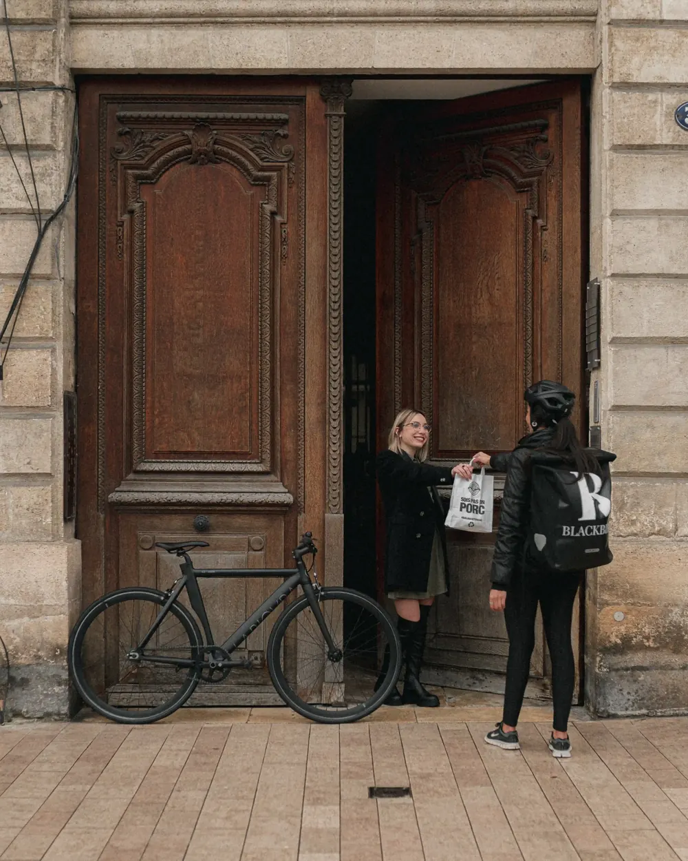 Une livreuse Blackbird remet une commande devant une porte cochère bordelaise, un vélo appuyé contre la façade